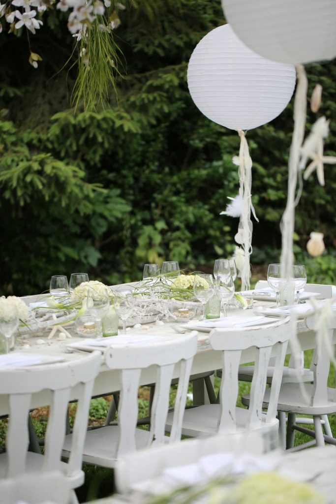 Elegantly set long white tables and chairs at an outdoor event planned by Rose Petal Family Events, with lush greenery in the background."